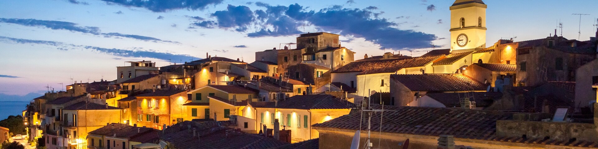 A coastal town at dusk with terracotta-roofed buildings, a prominent church tower, and a clear sky.
