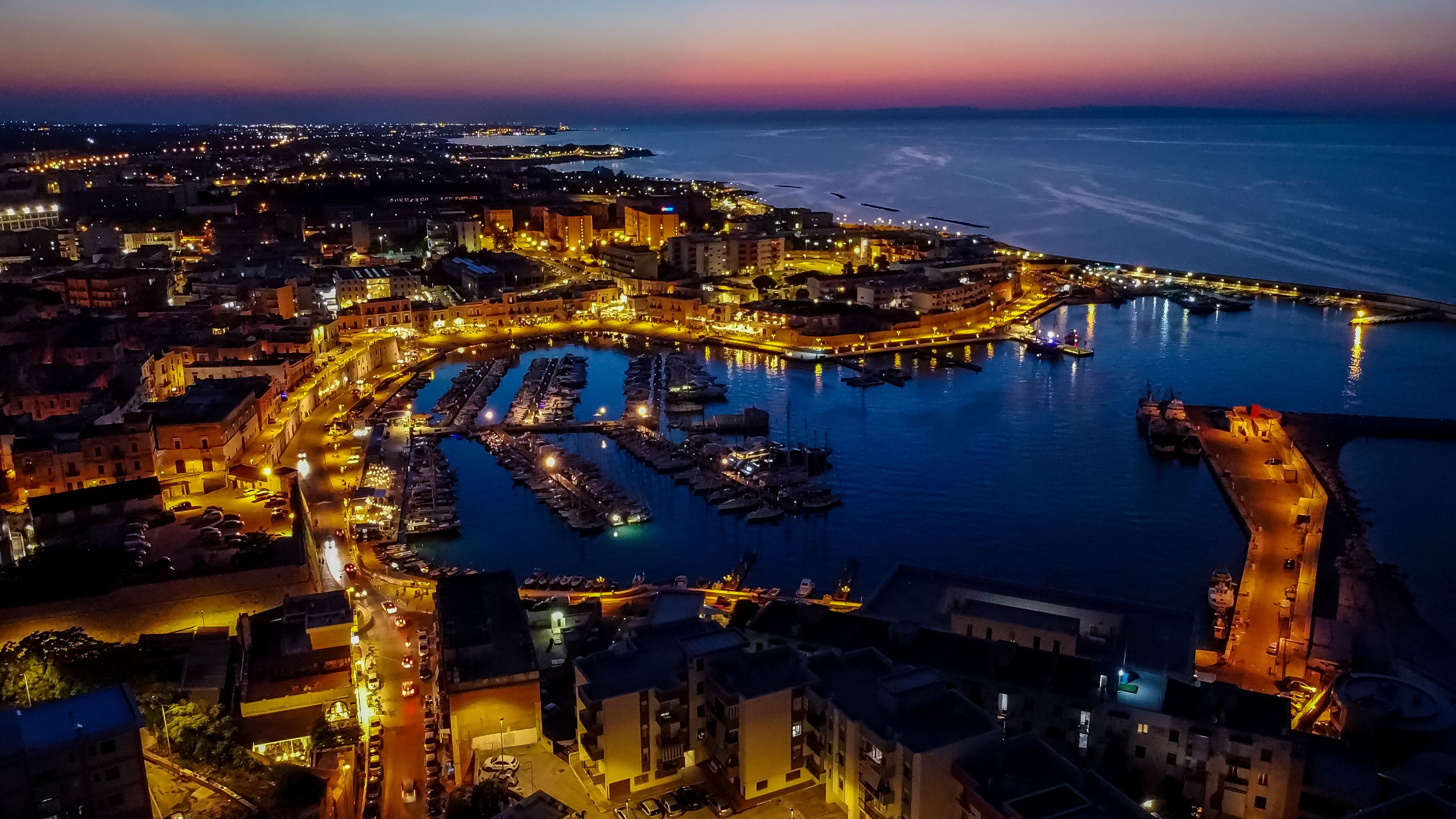A coastal cityscape at dusk with illuminated streets and a marina.