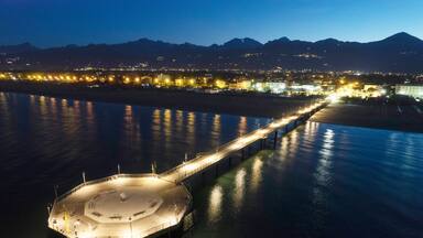 A waterfront pier extending into calm waters, surrounded by a cityscape with illuminated buildings and mountains in the background.