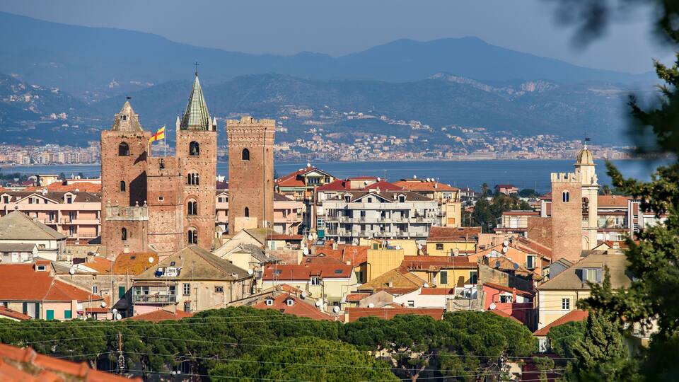 A coastal cityscape with prominent historical buildings, including a large church with twin towers and a bell tower, set against a backdrop of mountains and a clear blue sea.