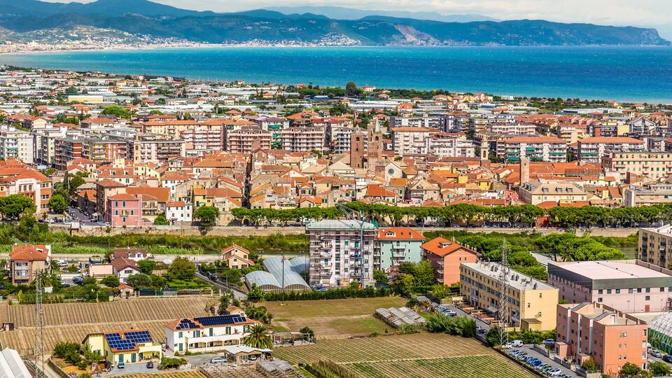A coastal cityscape with a mix of residential buildings, a prominent church, and a view of the sea and mountains in the background.