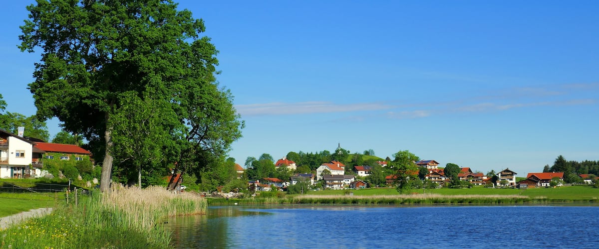 Blick vom Weg entlang Bad Soier See auf Bayersoien mit Ufer, Baum und Dorf unter blauem Himmel