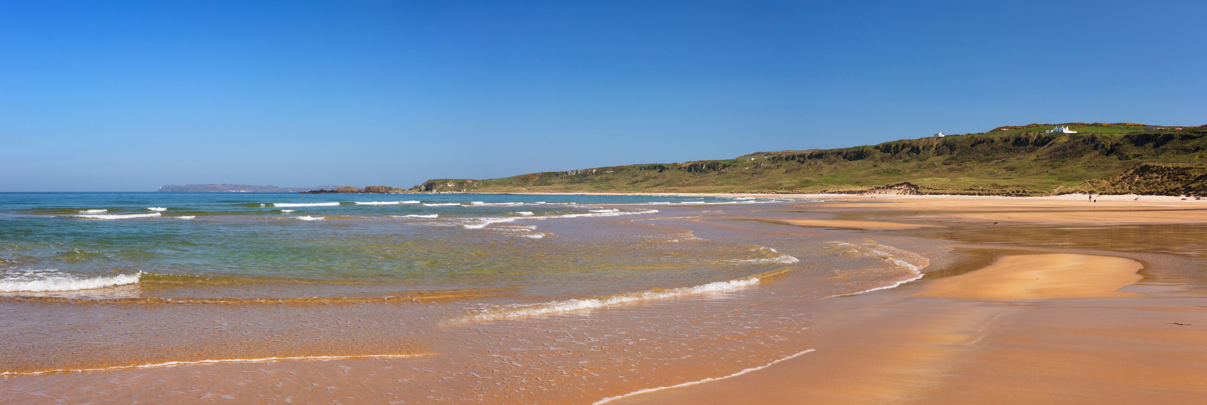 A sandy beach with gentle waves, a clear blue sky, and a grassy hill with a white building in the distance.
