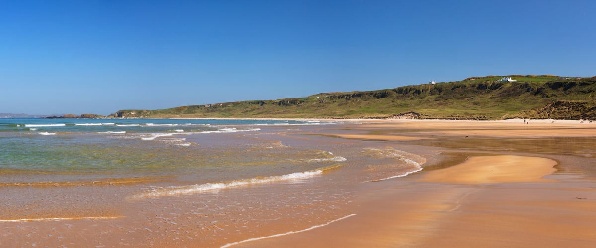 A sandy beach with gentle waves, a clear blue sky, and a grassy hill with a white building in the distance.