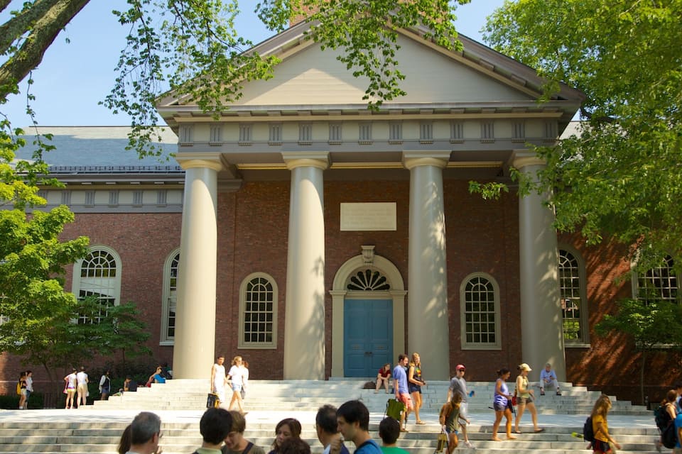 A classical building with columns and a blue door, surrounded by trees and people walking.