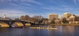 A river with a bridge, rowing team, and buildings in the background.