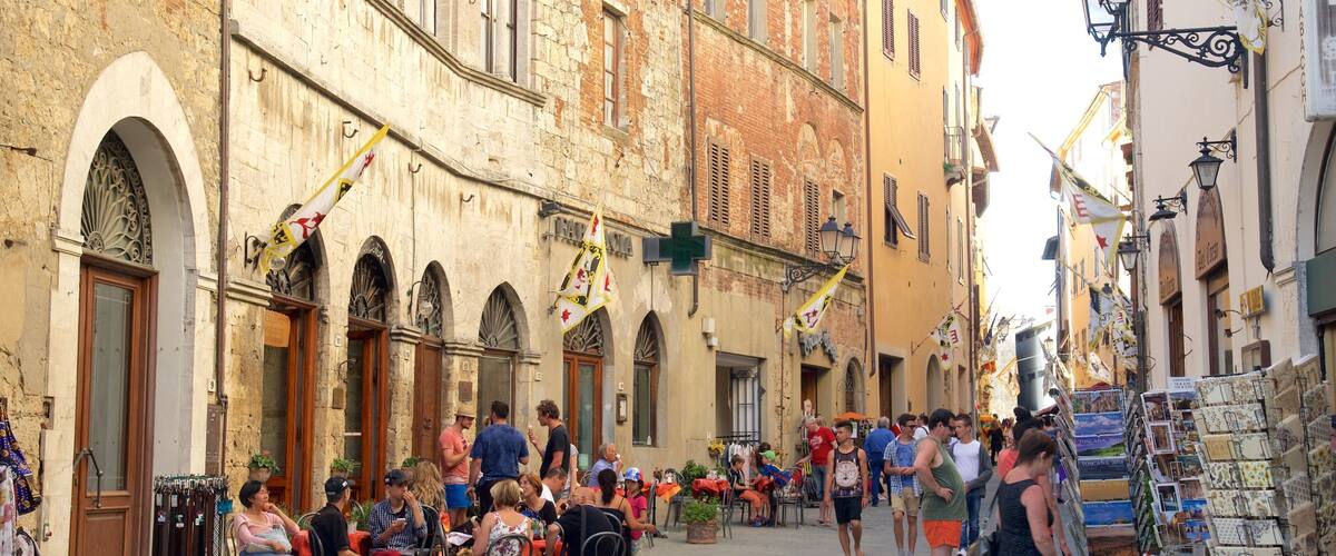 A crowded street with people sitting at tables, a vendor selling postcards, and a building with arched windows.