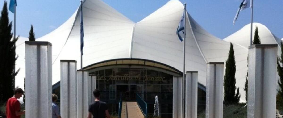 A modern architectural structure with a unique curved roof, surrounded by tall columns and a wooden boardwalk.
