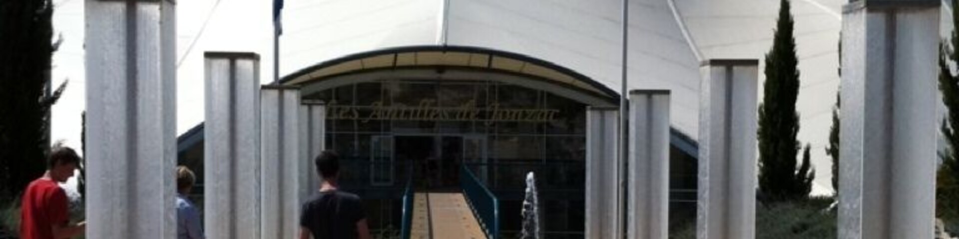 A modern architectural structure with a unique curved roof, surrounded by tall columns and a wooden boardwalk.