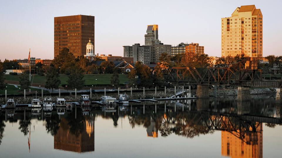 A city skyline with reflections in a calm body of water, including a clock tower, modern buildings, and a bridge.