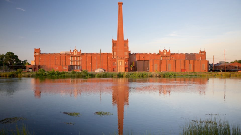 A large brick building with a tall chimney, reflecting in a calm body of water.