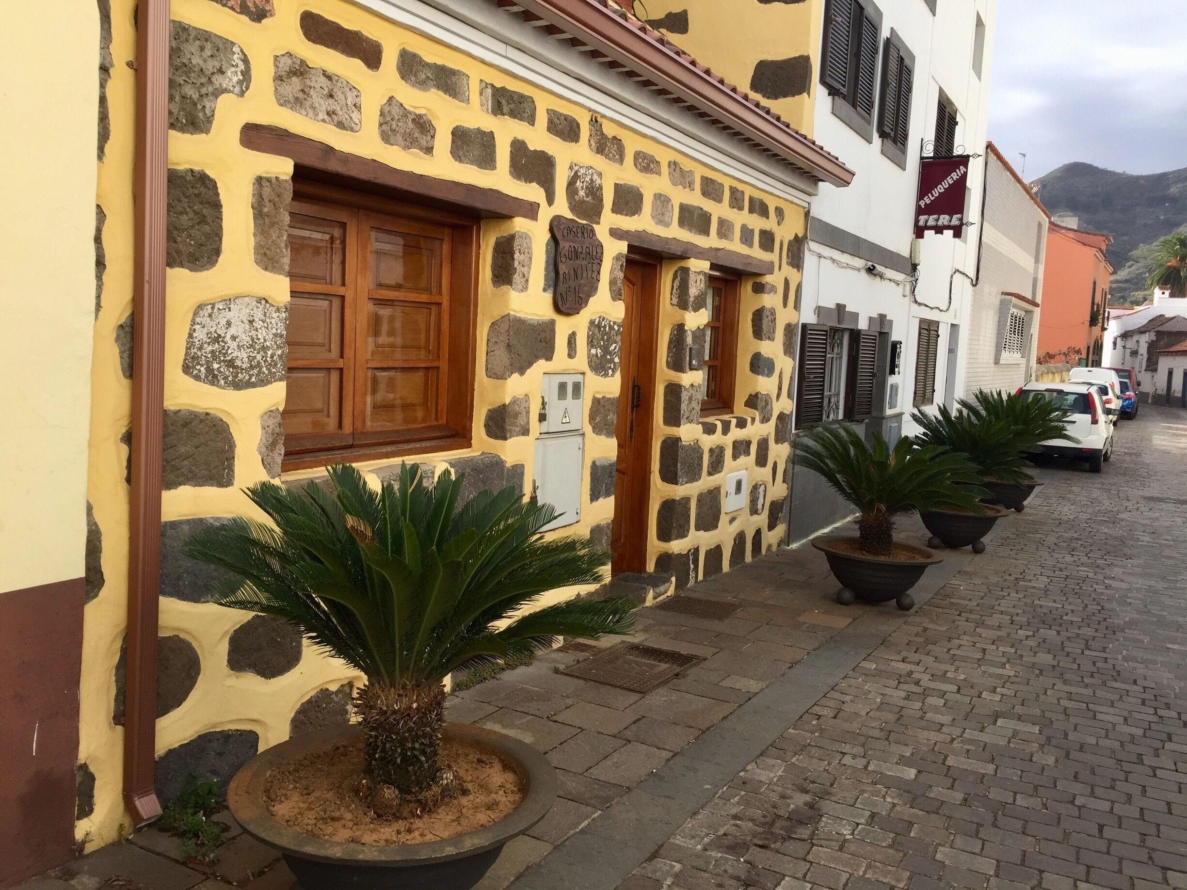 A cobblestone street with a yellow building featuring stonework and wooden shutters. There are potted plants and parked cars along the street.