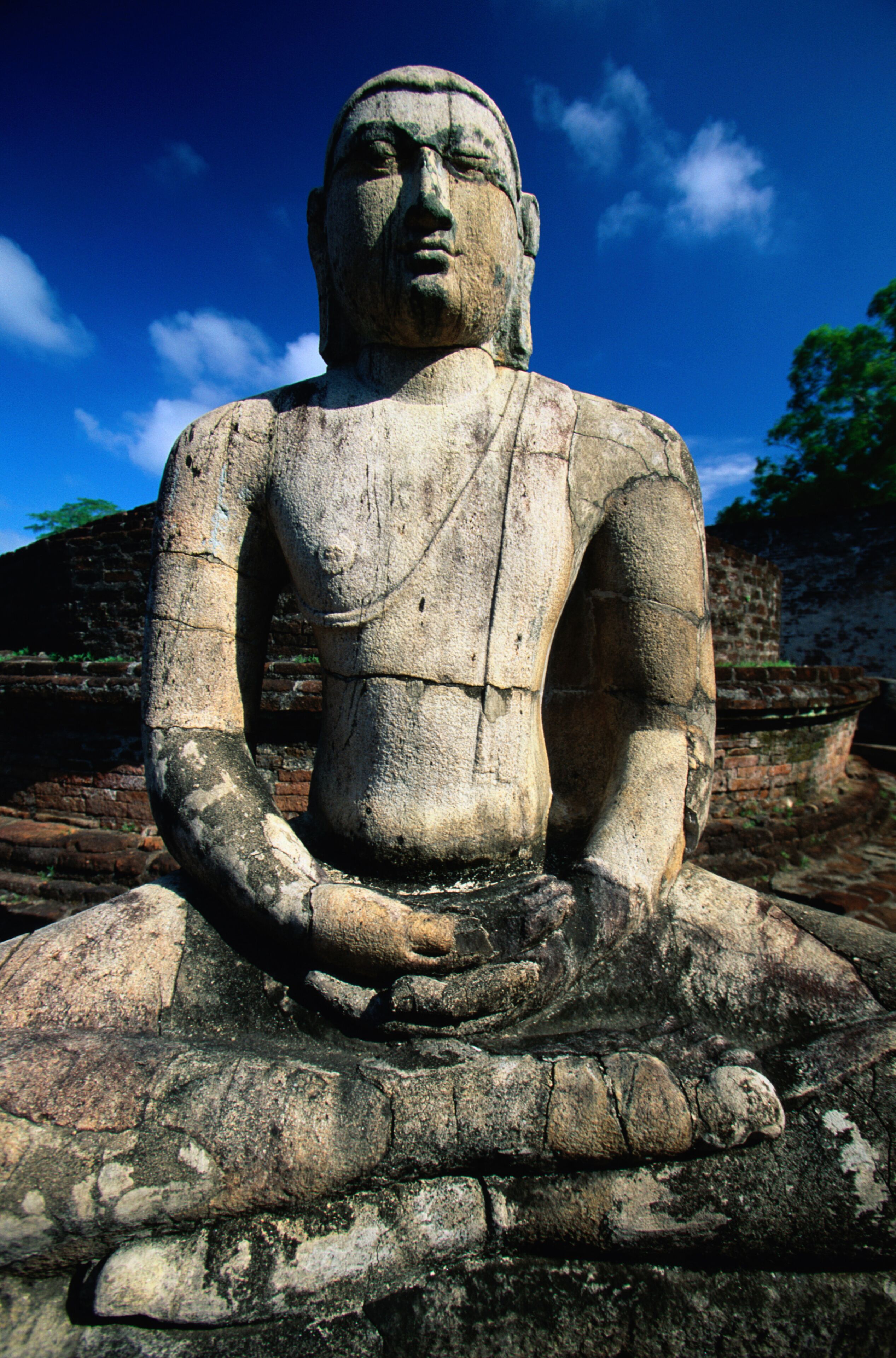 A large stone Buddha statue in a meditative pose, surrounded by ancient brick structures.