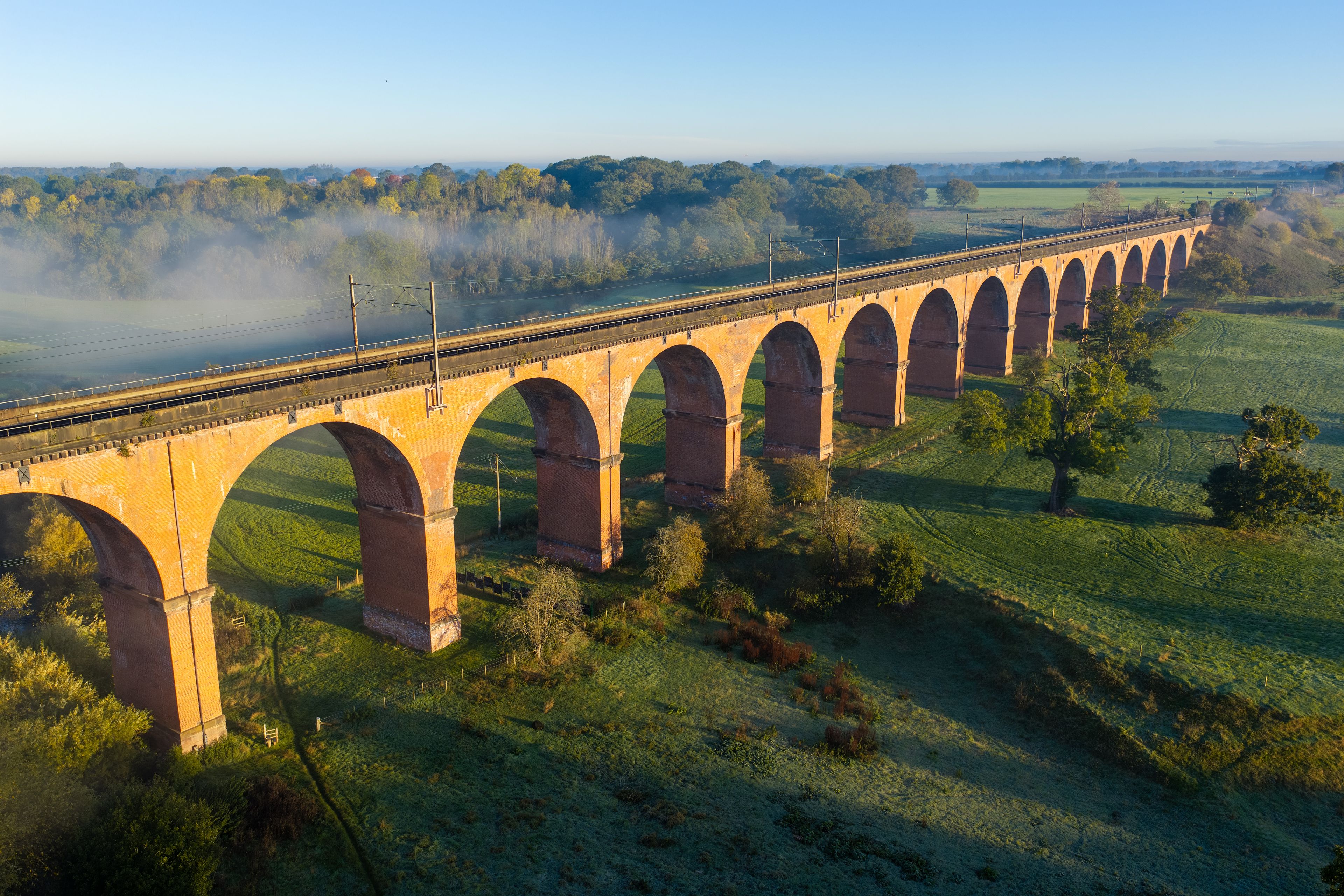 A long viaduct with multiple arches spanning a river, surrounded by green fields and trees.