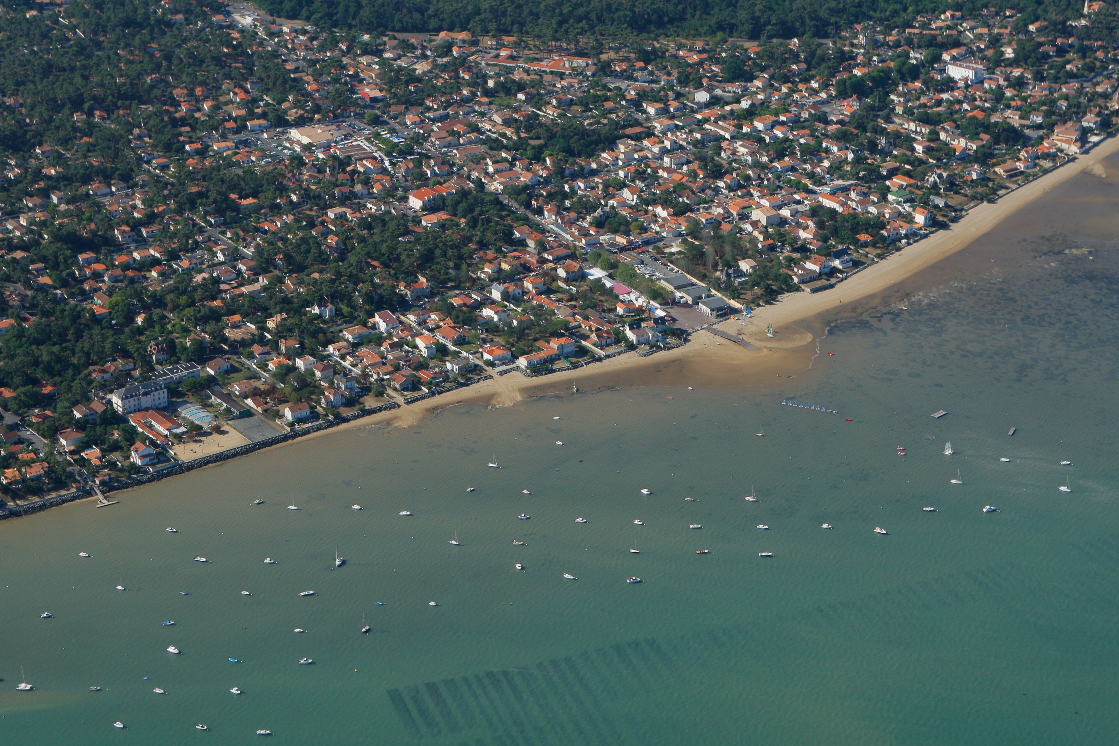 An aerial view of a coastal town with a beach, numerous boats, and dense residential areas.