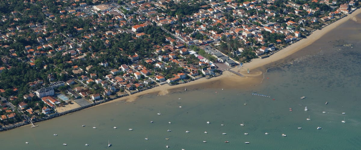An aerial view of a coastal town with a beach, numerous boats, and dense residential areas.
