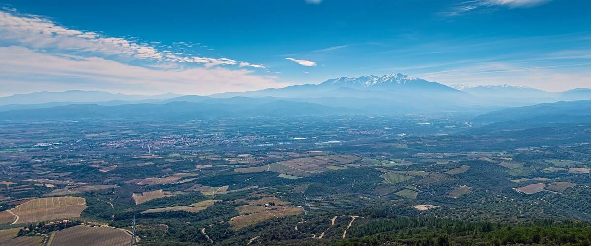 A mountainous landscape with snow-capped peaks, a valley with agricultural fields, and a clear blue sky.