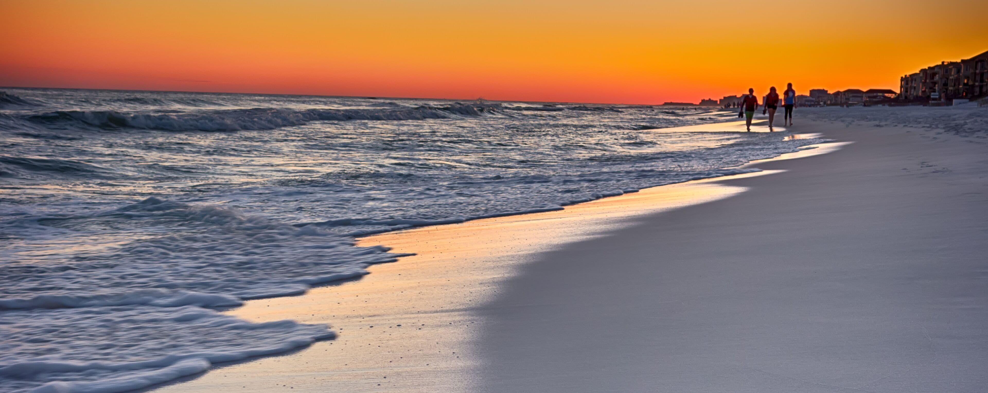 A beach at sunset with waves crashing onto the shore and people walking along the beach.
