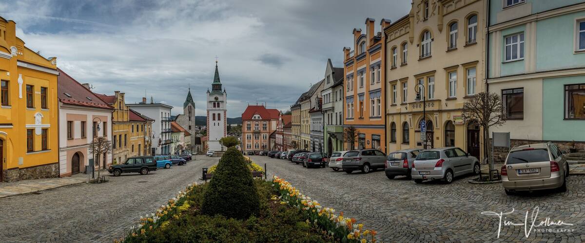 A cobblestone street lined with colorful buildings, a church with a green spire, and parked cars.