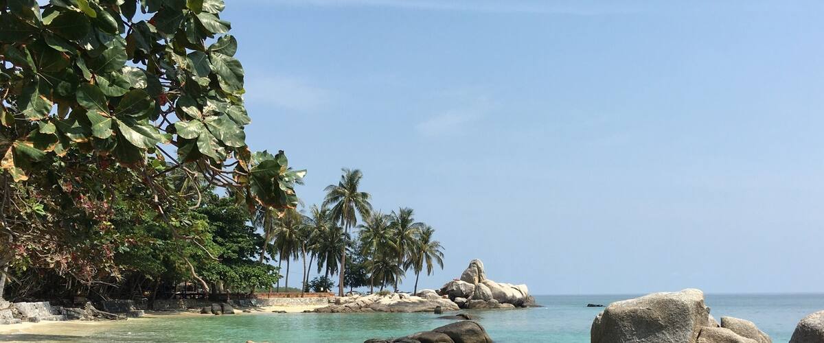 A tropical beach with large rocks, clear turquoise water, and palm trees.