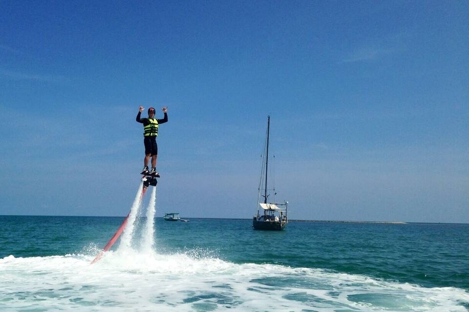 A person jet skiing on a water jetpack, with a sailboat in the background.
