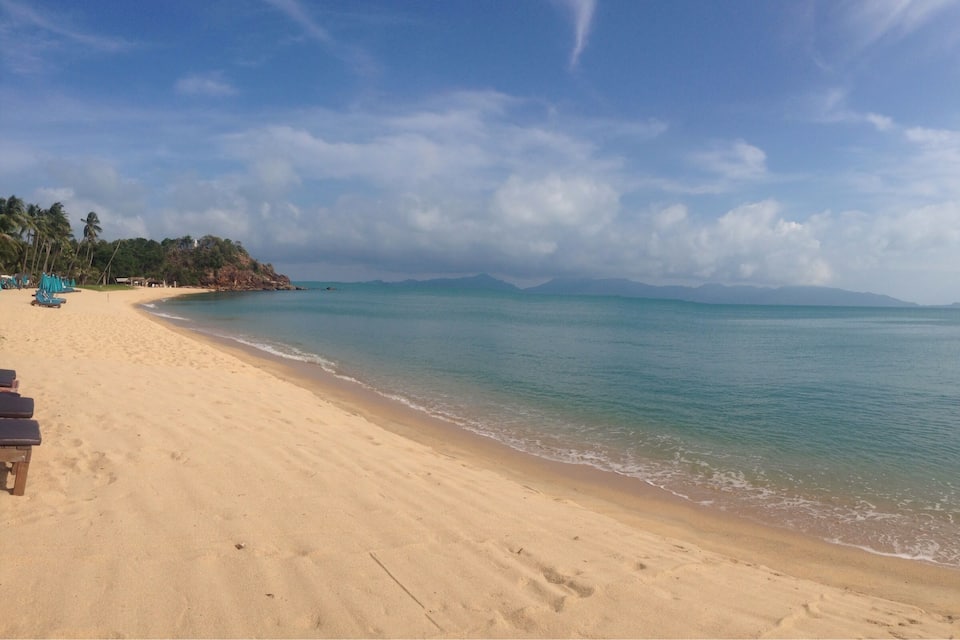 Une plage de sable fin, une mer calme, des palmiers et des montagnes à l’horizon.