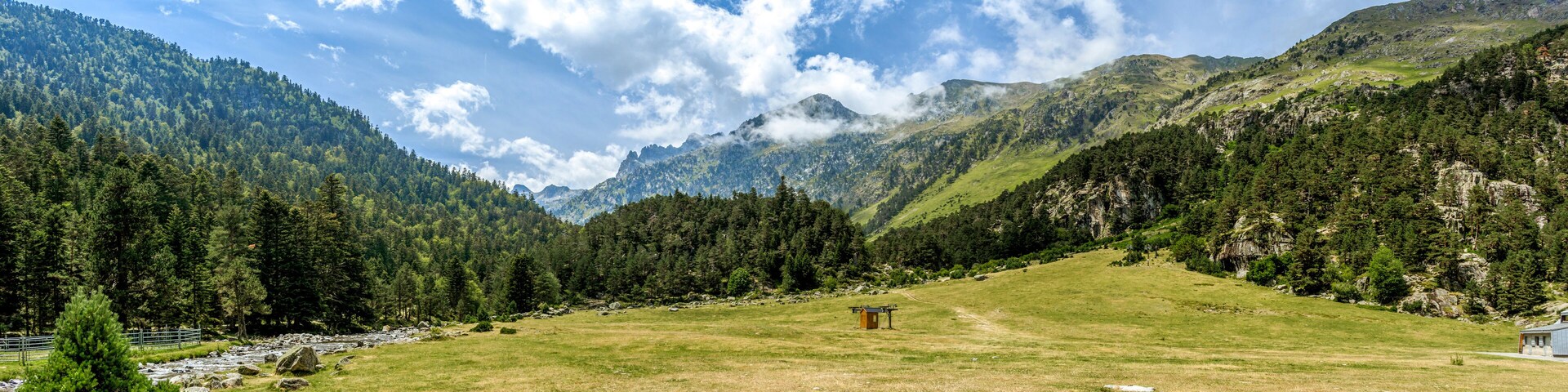 A mountainous landscape with a grassy field, a small building, and dense forests.