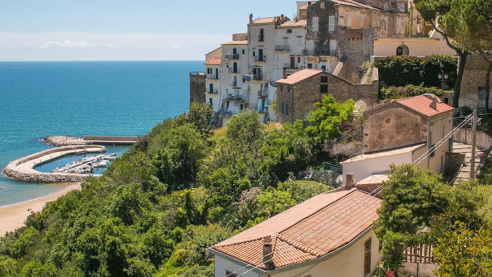A coastal town with terracotta-roofed buildings, a marina, and a beach.