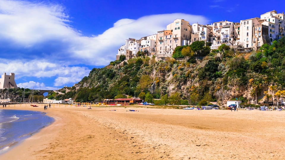 A beach with a castle on a cliff, a boat on the water, and people on the sand.