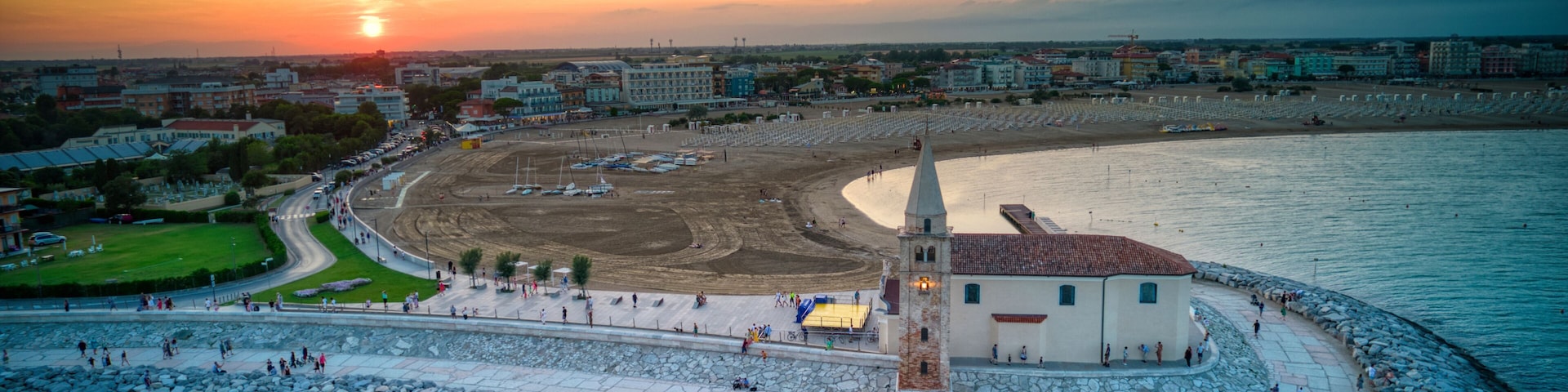 A coastal cityscape with a prominent lighthouse, a sandy beach, and a rocky breakwater.