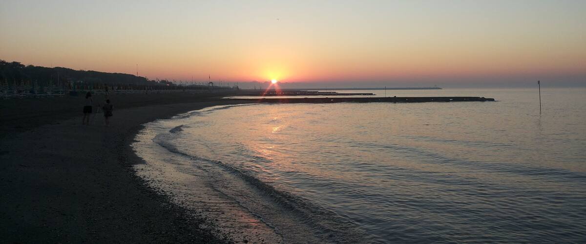 A beach at sunset with the sun low on the horizon, waves gently lapping the shore, and a few people walking along the beach.