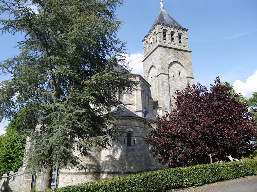 A historic stone church with a tall bell tower, surrounded by lush greenery and trees.