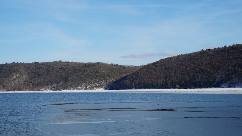 A frozen lake with snow-covered banks and a forested hill in the background.