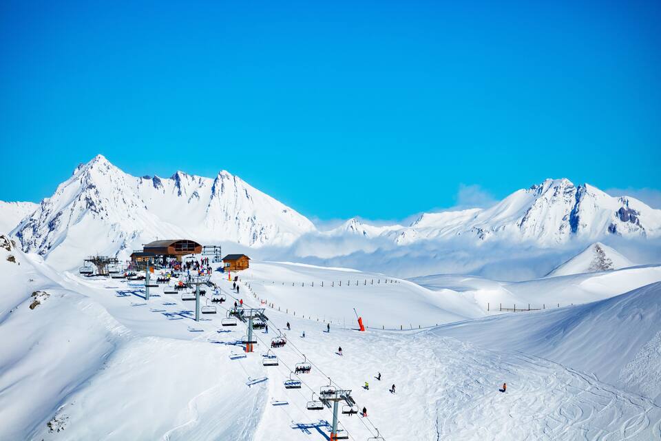 Una pista da sci innevata con una seggiovia, un rifugio e montagne sullo sfondo.