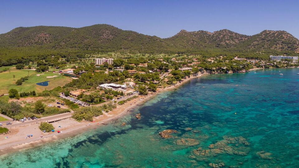 A coastal town with a beach, clear blue waters, and green hills in the background.