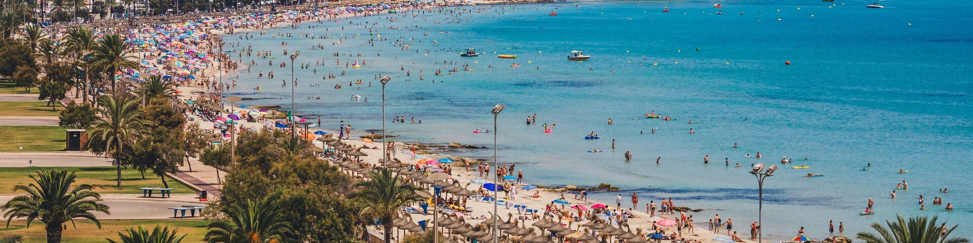 Una playa concurrida, con aguas azules cristalinas, palmeras y montañas al fondo.