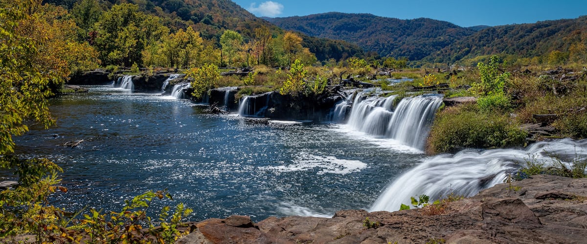 A multi-tiered waterfall in a forested valley with mountains in the background.