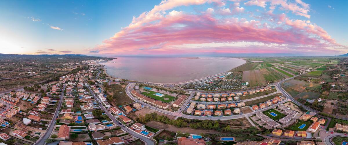 An aerial view of a coastal residential area with a large body of water, a curved road, and surrounding fields.