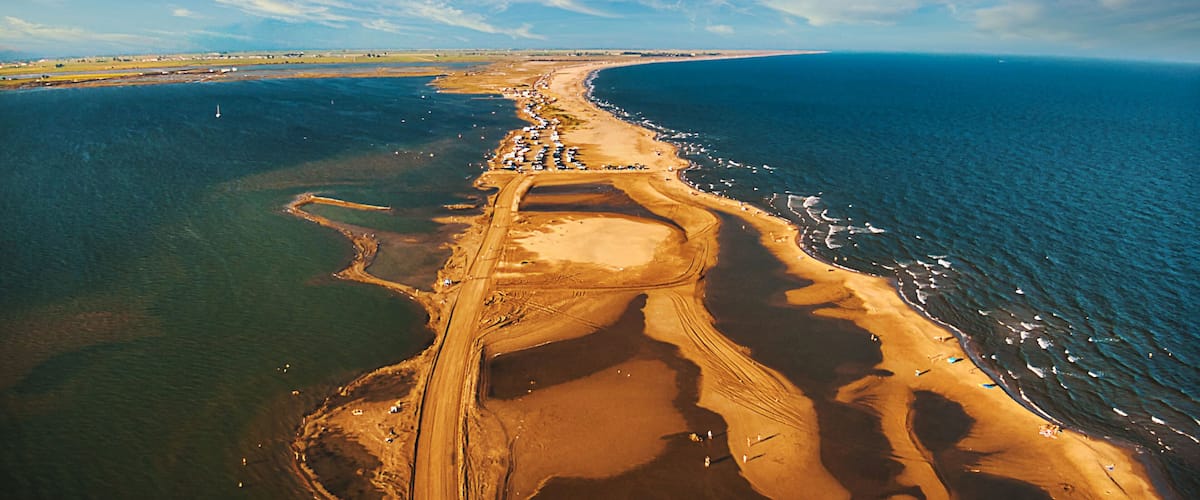 An aerial view of a coastal area with a long sandy beach, a clear blue sky, and a calm sea.
