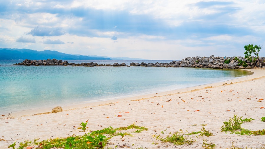 Una playa con agua turquesa cristalina, arena blanca y un rompeolas rocoso.