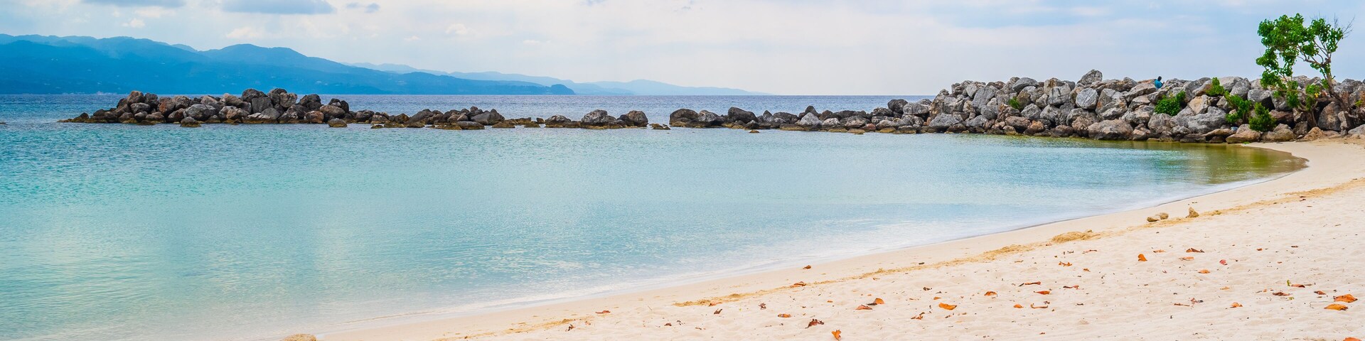 Una playa con agua turquesa cristalina, arena blanca y un rompeolas rocoso.