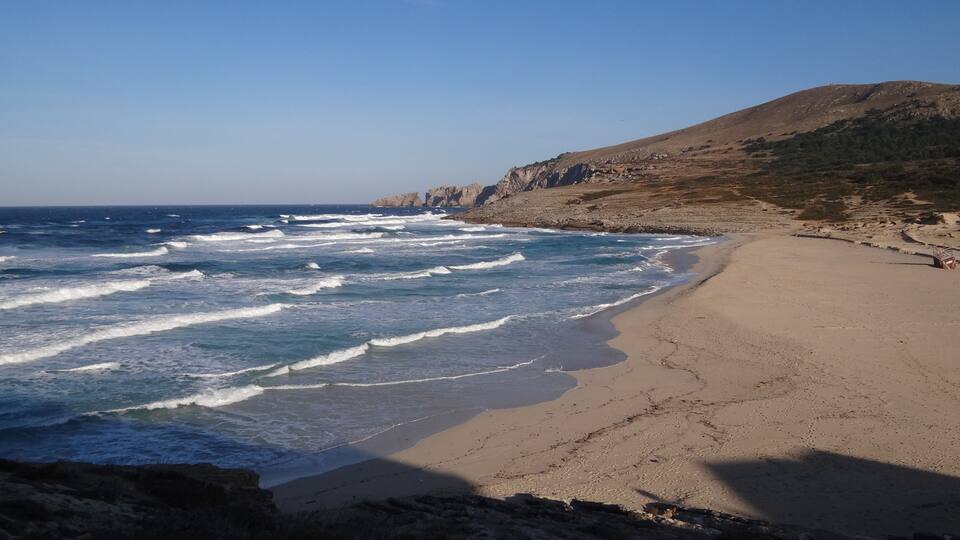 A beach with waves crashing onto the shore, a rocky cliff, and a clear blue sky.