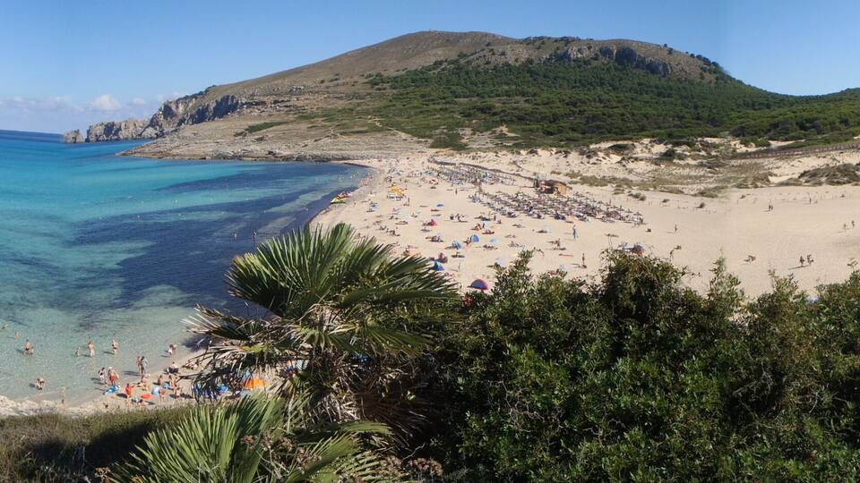 A coastal beach with clear turquoise waters, a sandy shore, and a mountainous backdrop.