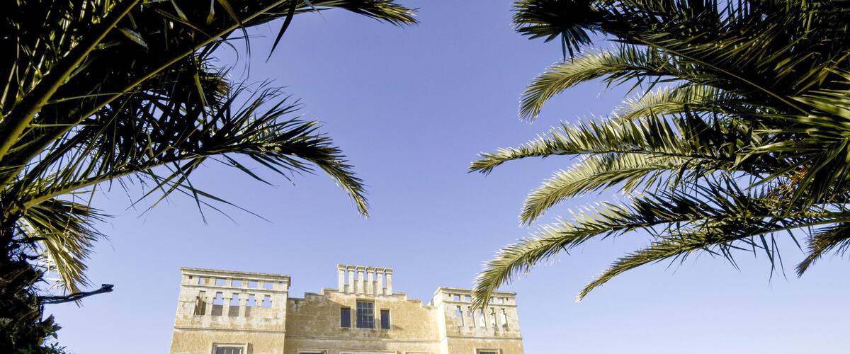 A historic stone building with arched windows and a central entrance, surrounded by stone walls and palm trees.