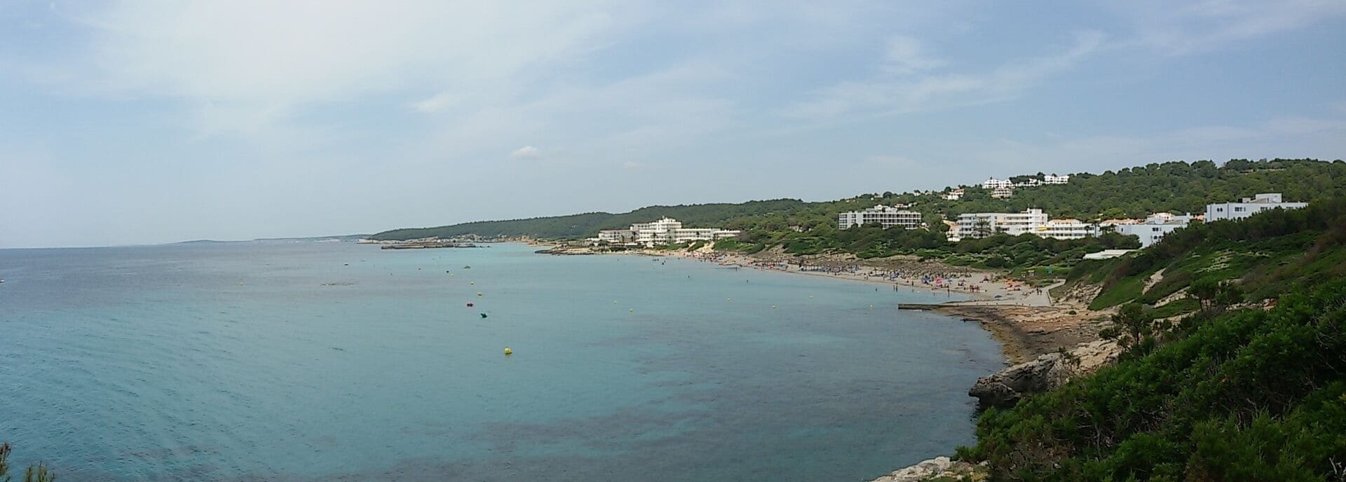 A coastal landscape with a clear blue sea, sandy beach, and buildings on a hillside.
