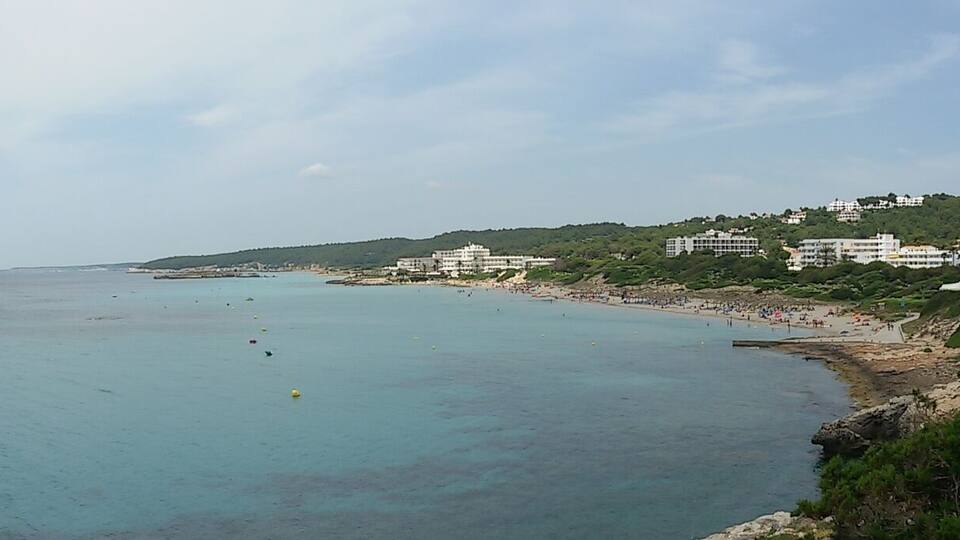 A coastal landscape with a clear blue sea, sandy beach, and buildings on a hillside.