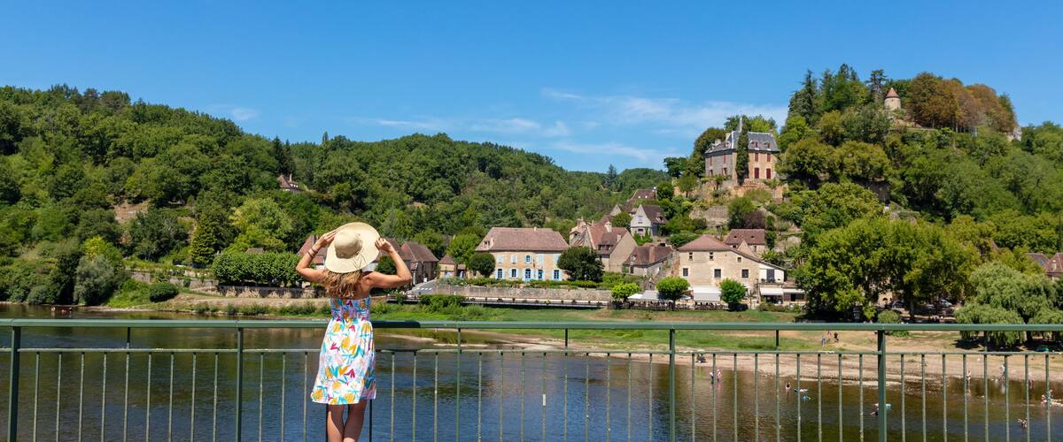 A woman in a dress and hat standing by a river, overlooking a town with historic buildings and a castle on a hill.