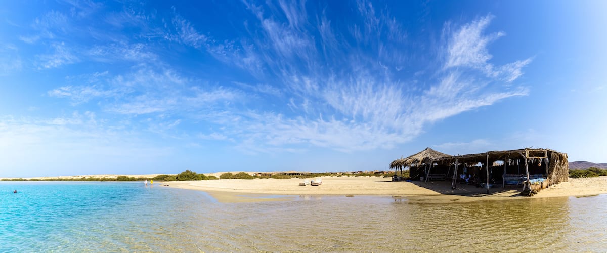 A beach with clear blue water, a thatched-roof hut, and distant dunes.