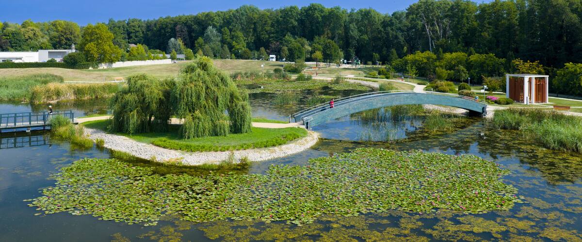 A park with a curved bridge over a pond, surrounded by trees and a small building.