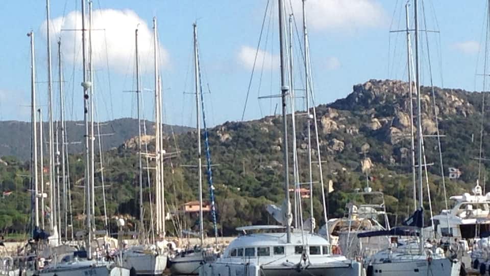 A marina with multiple sailboats docked, surrounded by a mountainous landscape.